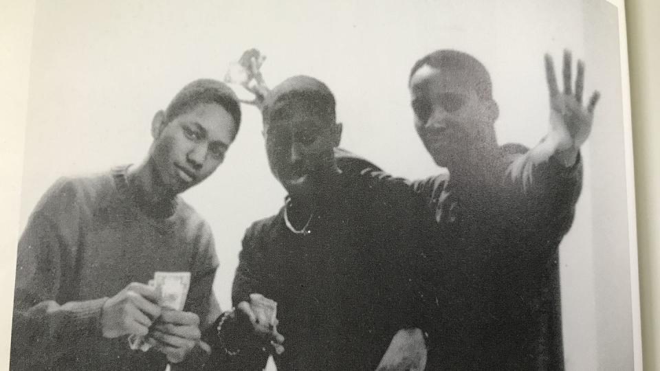 tupac shakur and two friends post for a photo, tupac is in the middle and holds some cash in one hand