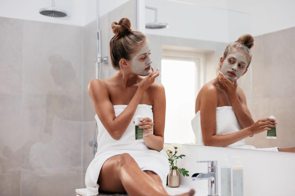 Young woman sitting on the sink counter applying a face mask while looking in a mirror.