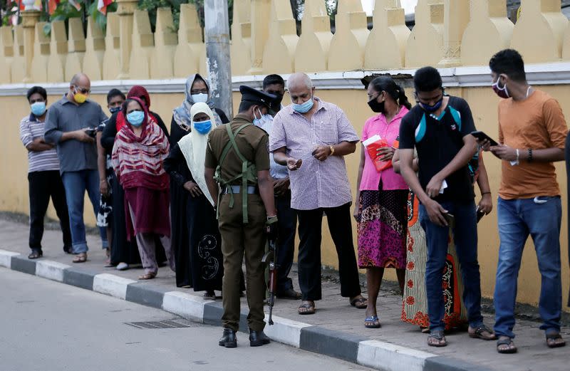 People wearing protective masks wait in line to cast their votes outside a polling station while a police officer checks thier polling cards during the country's parliamentary election in Colombo