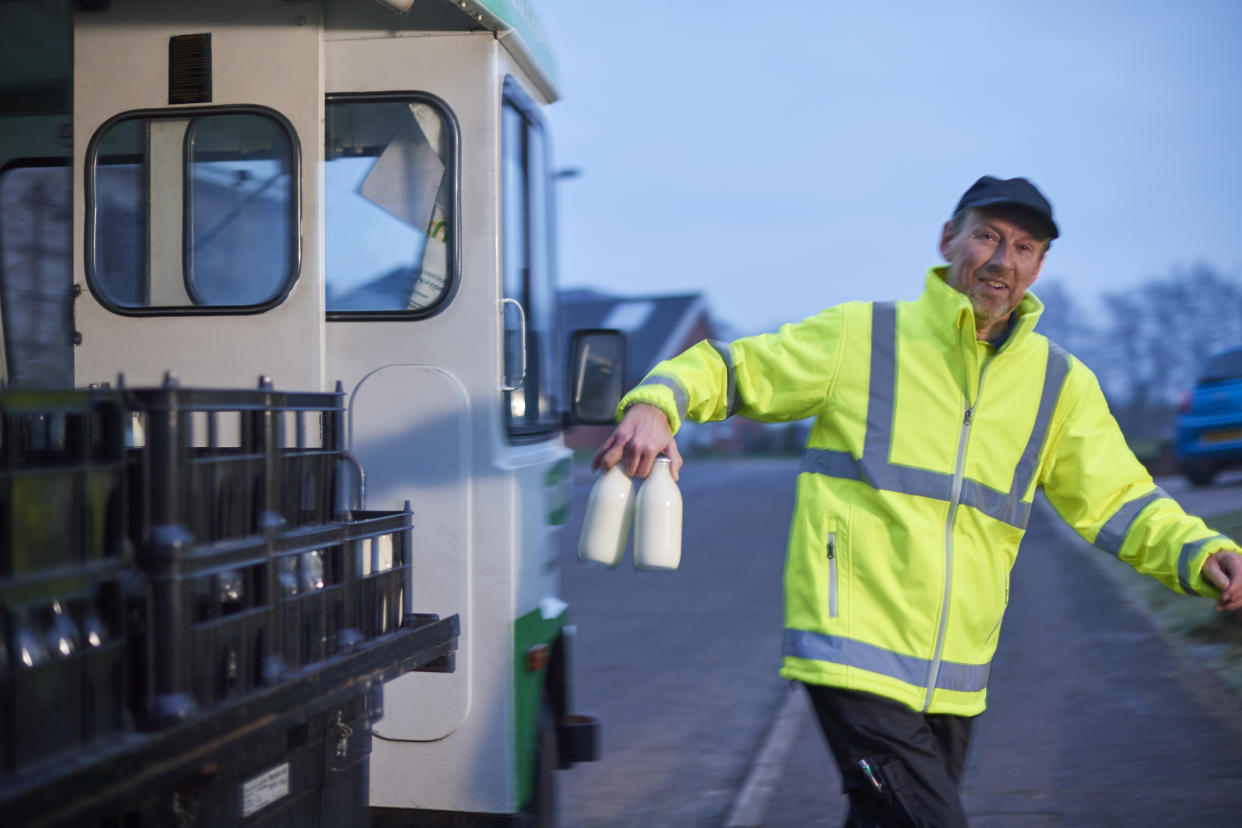 Milkman wearing fluorescent yellow jacket, smiling and walking from his milk float with milk bottles in hand on urban street in morning light
