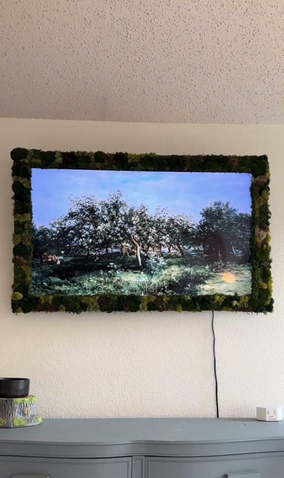 A TV screen hanging on wall displaying a landscape scene, framed with decorative moss