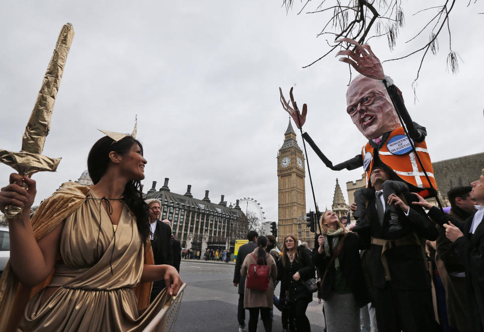 Lawyers, one dressed as Justice, left, and one carrying a puppet depicting Chris Grayling, Britain's Justice Secretary march to protest against legal aid cuts, across from the Houses of Parliament in central London, Friday, March 7, 2014. The protest coincides with a nationwide demonstration of non-attendance of lawyers which will affect hundreds of cases across the country. (AP Photo/Lefteris Pitarakis)