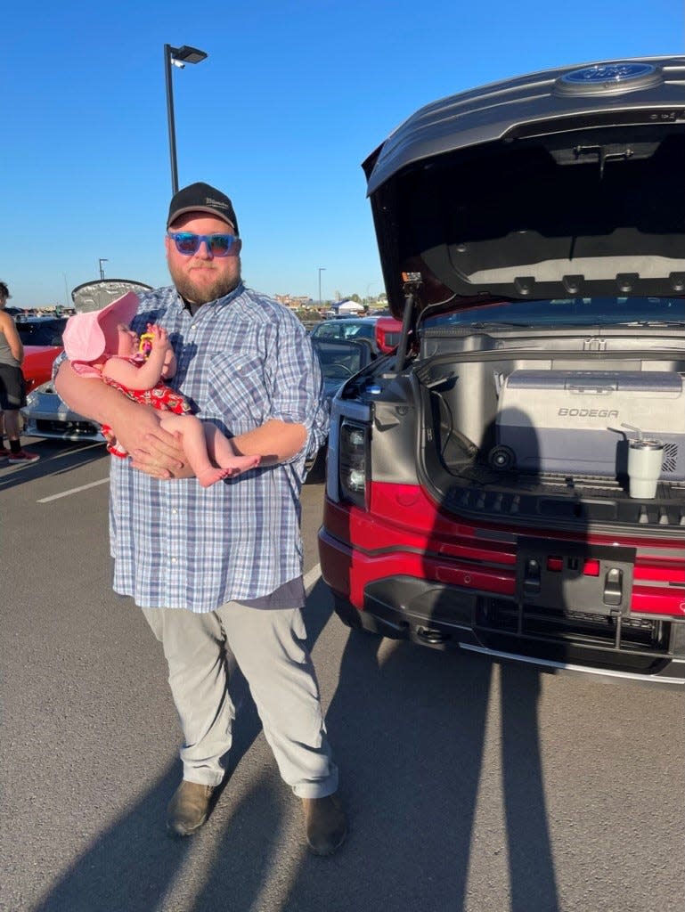 Mac Jaehnert of Richland, Washington, holds his daughter, MacKenzie, with an electric cooler plugged into the front trunk of his all-electric Ford F-150 Lightning. The cooler holds beverages and baby bottles at a Cars & Caffeine event in Pasco, Washington, on Aug. 13, 2022.