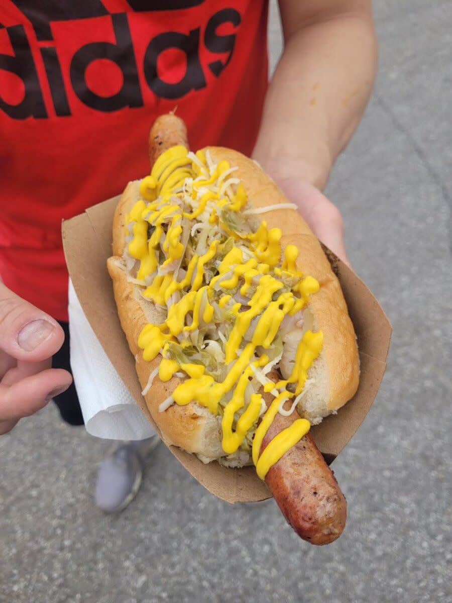 Man wearing a red shirt holding a KC Frank in a cardboard boat from Wiener Kitchen, Kansas City, his shoes and concrete are blurred in the background