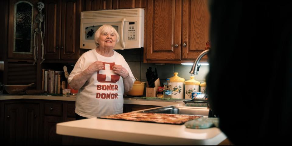 June Squibb in Hubie Halloween, stands in a kitchen and proudly shows a white T-shirt with a cross symbol and the text "Stand dispenser"