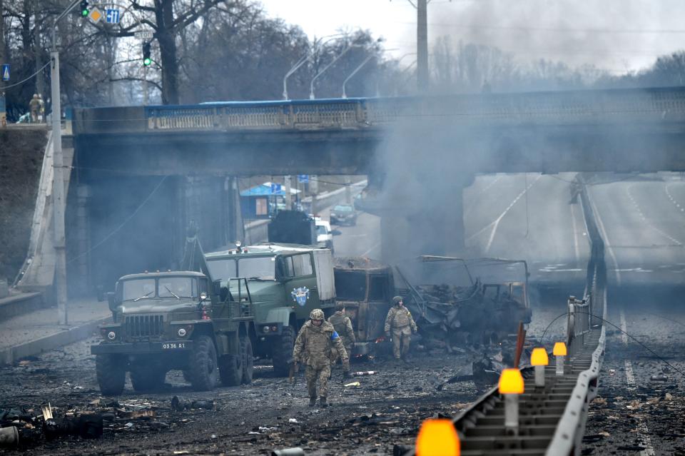 Ukrainian service members walk next to military vehicles amid smoke and debris.