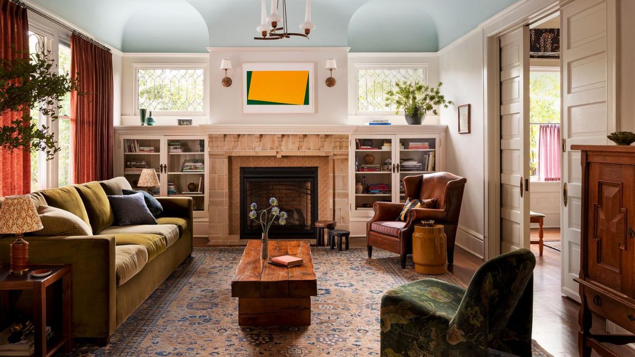 ocher velvet sofa in front of curtained windows in a rust color, bookshelves flank a fireplace with small leaded windows above, leather armchair, slipper chair, cocktail table, turkish rug, vintage chandelier