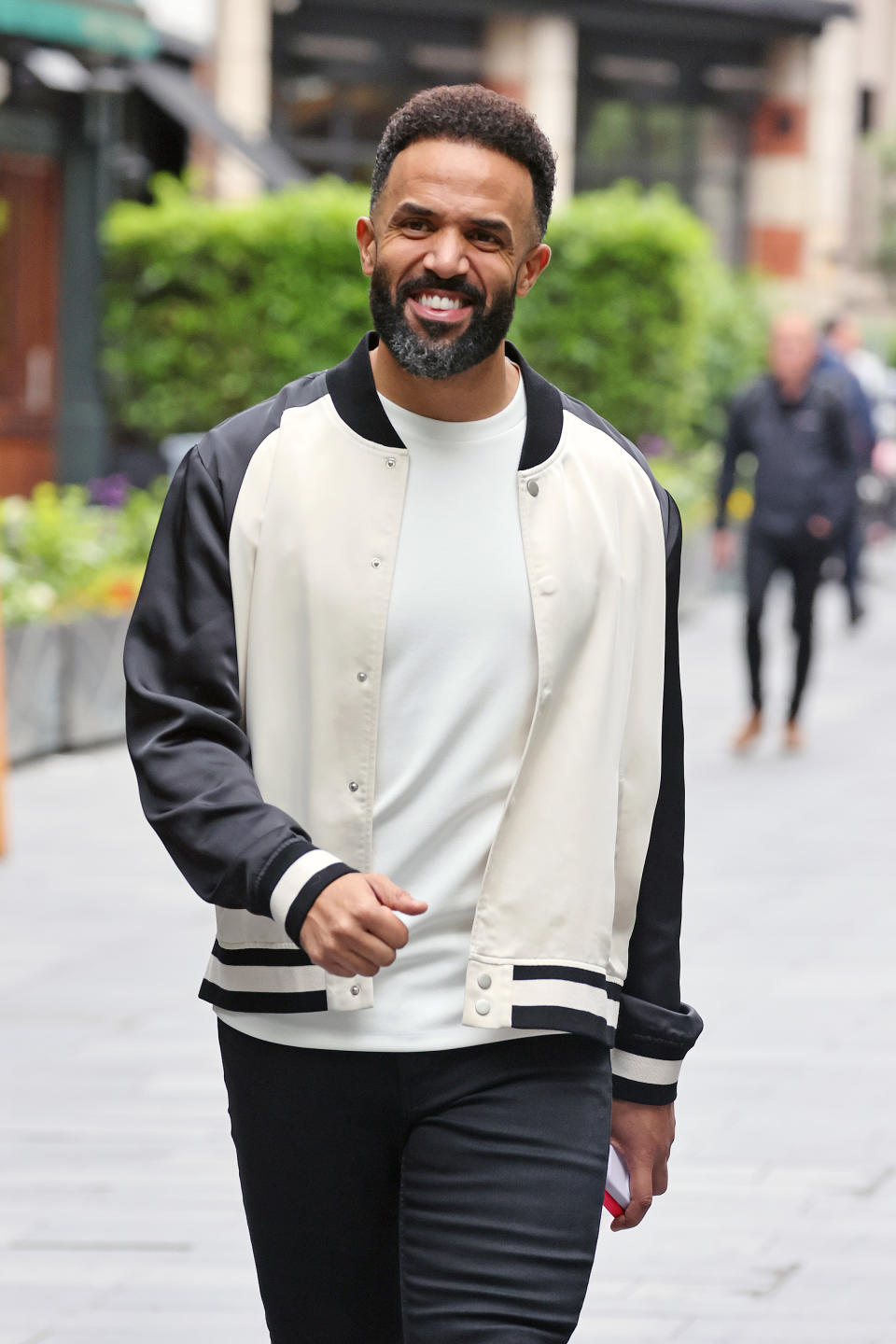 Craig David smiles while walking outside wearing a white t-shirt, black jeans, and a black-and-white varsity jacket