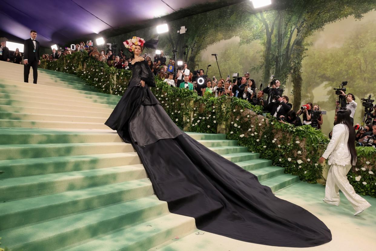 Zendaya walks up the red carpet of the 2024 Met Gala, wearing a flower headdress and a black gown that drapes down the steps behind her.