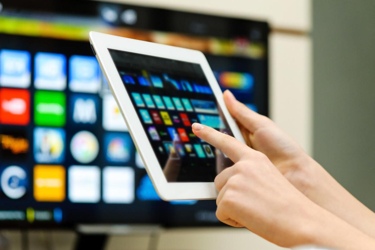 Focus on two female hands holding a tablet, choosing a channel for TV in the background with the blurred background of the TV and wall