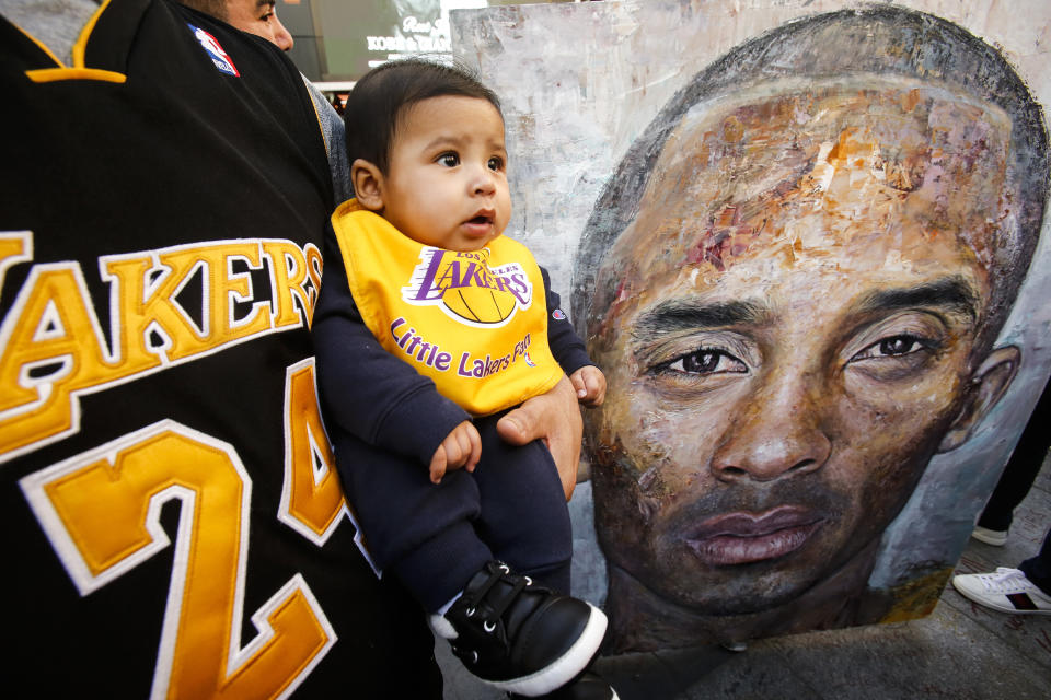 Juan Quijas holds his son Danny in front front of a painting of the late Kobe Bryant at a memorial near Staples Center prior to an NBA game between the Los Angeles Lakers and the Portland Trail Blazers, Friday, Jan. 31, 2020, in Los Angeles. Bryant, the 18-time NBA All-Star who won five championships and became one of the greatest basketball players of his generation during a 20-year career with the Los Angeles Lakers, died in a helicopter crash Sunday. (AP Photo/Ringo H.W. Chiu)