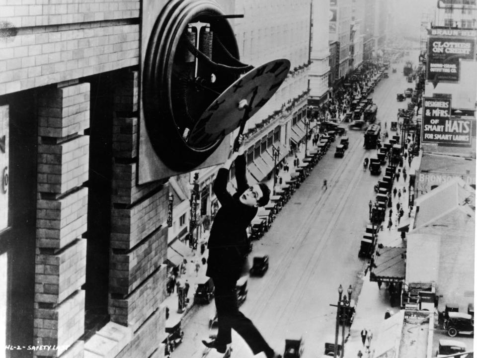 Harold Lloyd hangs from the arms of a broken clock on the side of a building high above a traffic-filled city street