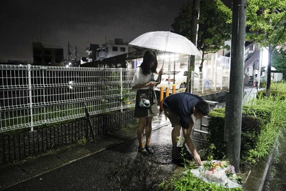A couple places flowers near the Kyoto Animation building, Friday, July 19, 2019, background left, that was destroyed in an arson attack in Kyoto, Japan. A man screaming "You die!" burst into an animation studio in Kyoto, doused it with a flammable liquid and set it on fire Thursday, killing several people in an attack that shocked the country and brought an outpouring of grief from anime fans. (AP Photo/Jae C. Hong)