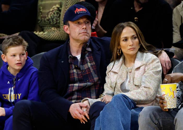 Jennifer Lopez sits courtside with Ben Affleck and his son Samuel at a basketball game. She wears a stylish jacket; Affleck wears a hat and casual outfit