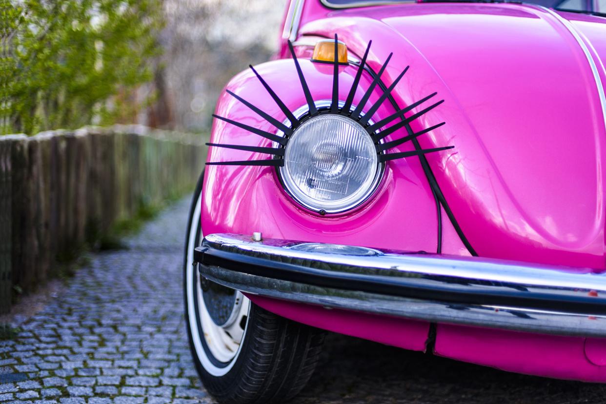 Closeup of front light with black eyelash of bright pink Volkswagen Type 1 Beetle beetle on a small cobblestone road with a blurred background of a fence and trees