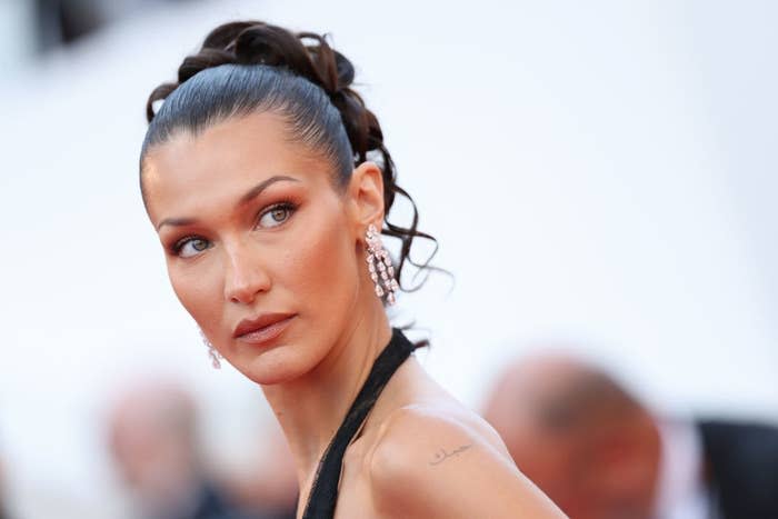 Bella Hadid on the red carpet, wearing elegant drop earrings and a black gown with her hair styled in an updo. She is looking to the side