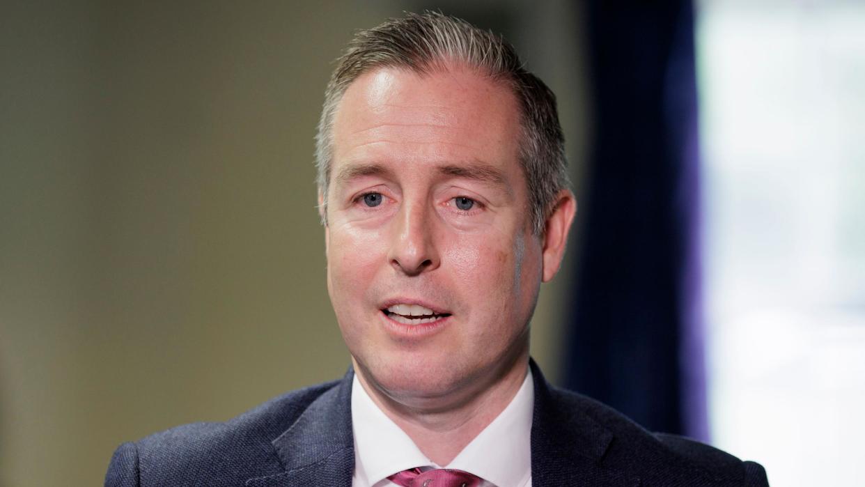 A man with dark grey hair is speaking to someone off-camera. He is wearing a dark blue suit, a white shirt and a crimson tie. 