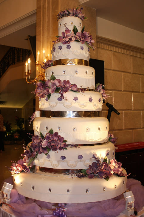 Tall, multi-tiered wedding cake decorated with purple flowers and small white lights, displayed indoors