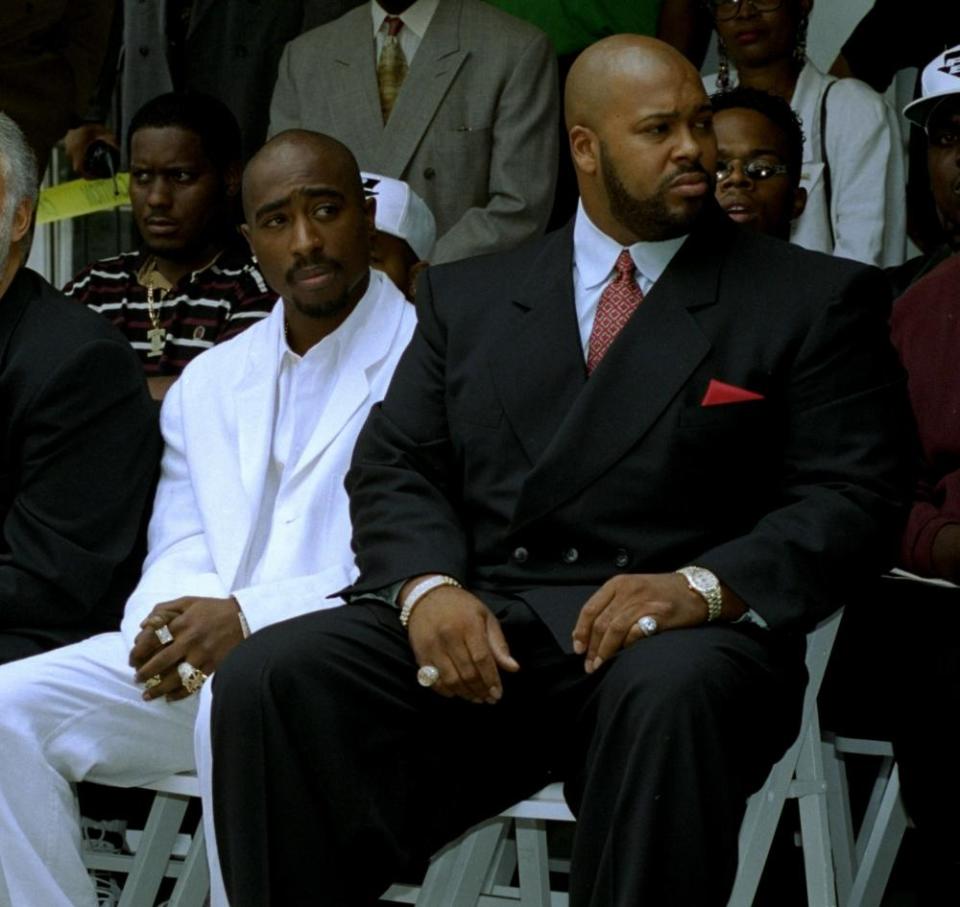 Seated on adjacent white folding chairs, amid rows of other people on white folding chairs, is a young Black man with a bald head and black mustache wearing a white suit, next to a much larger Black man, also with a bald head, who has a black beard and wears a black suit, light blue shirt, red tie, and red pocket square. The man’s hands rest on his thighs and showing two large pinky rings.