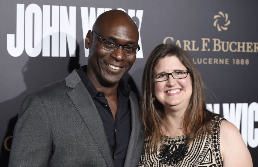 A bald man with glasses in a suit smiling and posing with a smiling brunet woman in a patterned dress