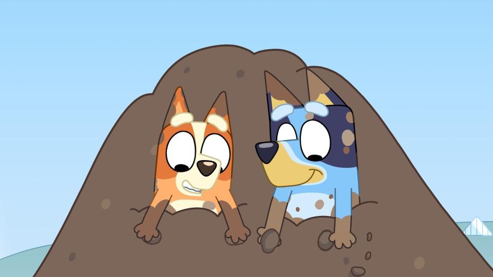 Animated characters Bluey and Bingo peek out from a dirt mound in the show "Bluey."
