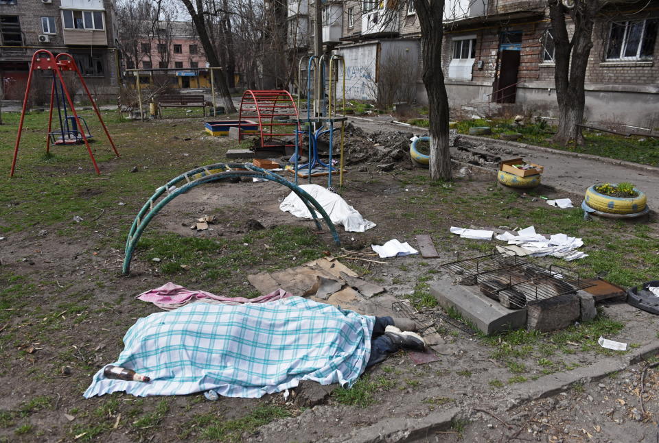 Two bodies covered with sheets lie in a children's playground, with a sheaf of papers strewn next to a grill.