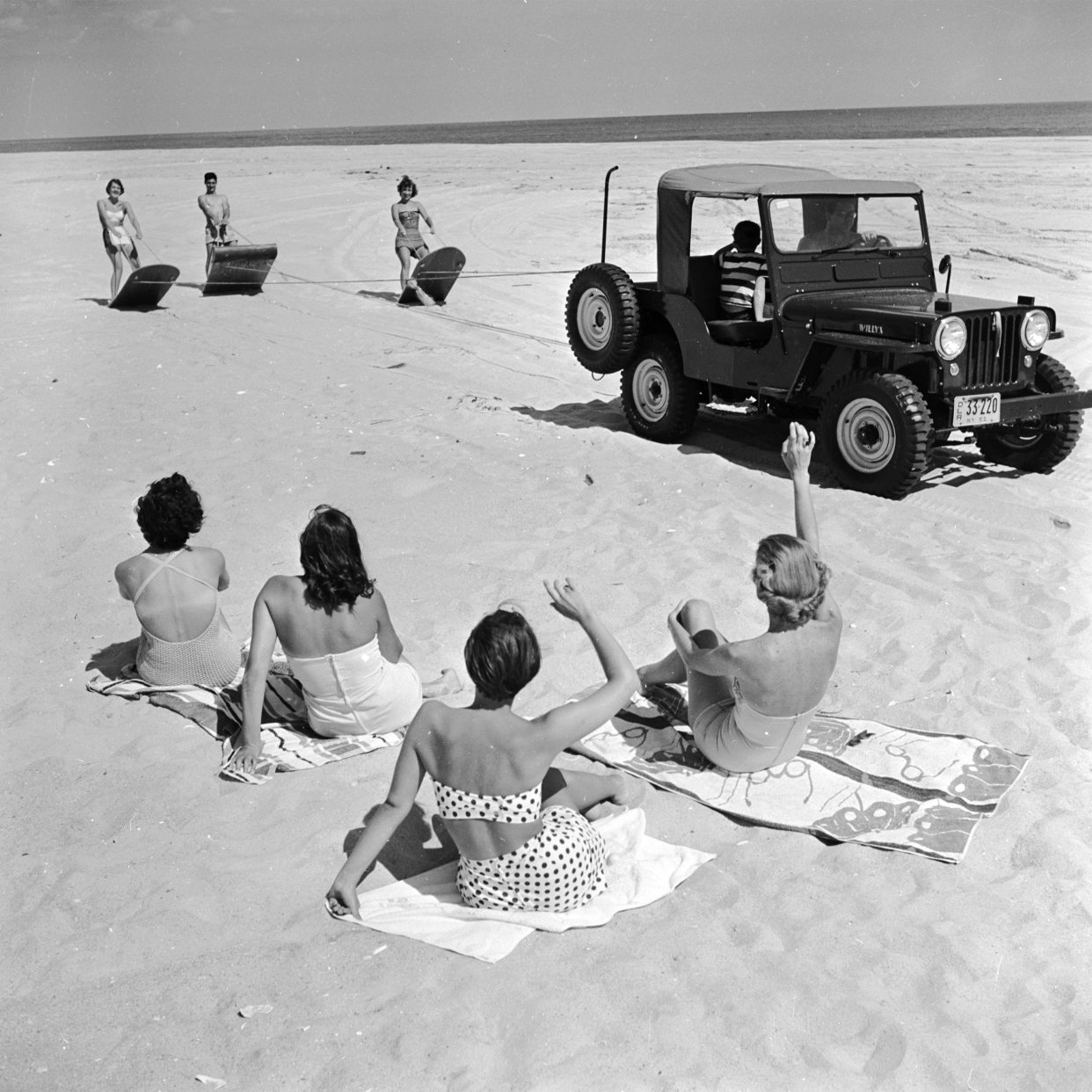 Sun bathers on a beach near Southampton, Long Island, USA, greet a trio of surfers pulled along the beach by an all-terrain vehicle.