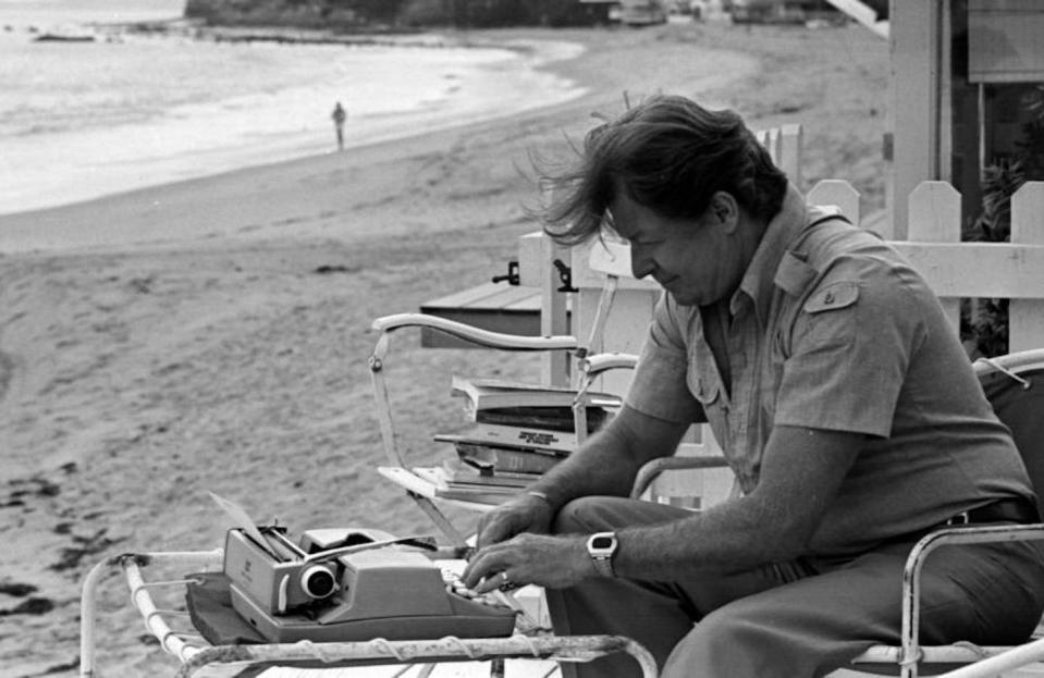 Black and white photo of middle-aged man sitting at a typewriter by the ocean.
