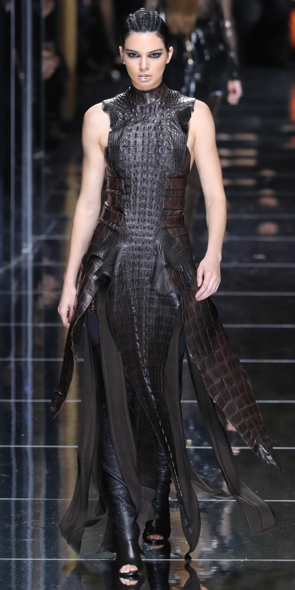 <p>Jenner looked gladiator chic at Balmain in a reptilian slit dress over leather leggings and peep toe boots, but the most surprising part of the ensemble was her makeup. The model looked almost unrecognizable in tight braids, black graphic liner, and a piece of gold jewelry on her lips.</p>