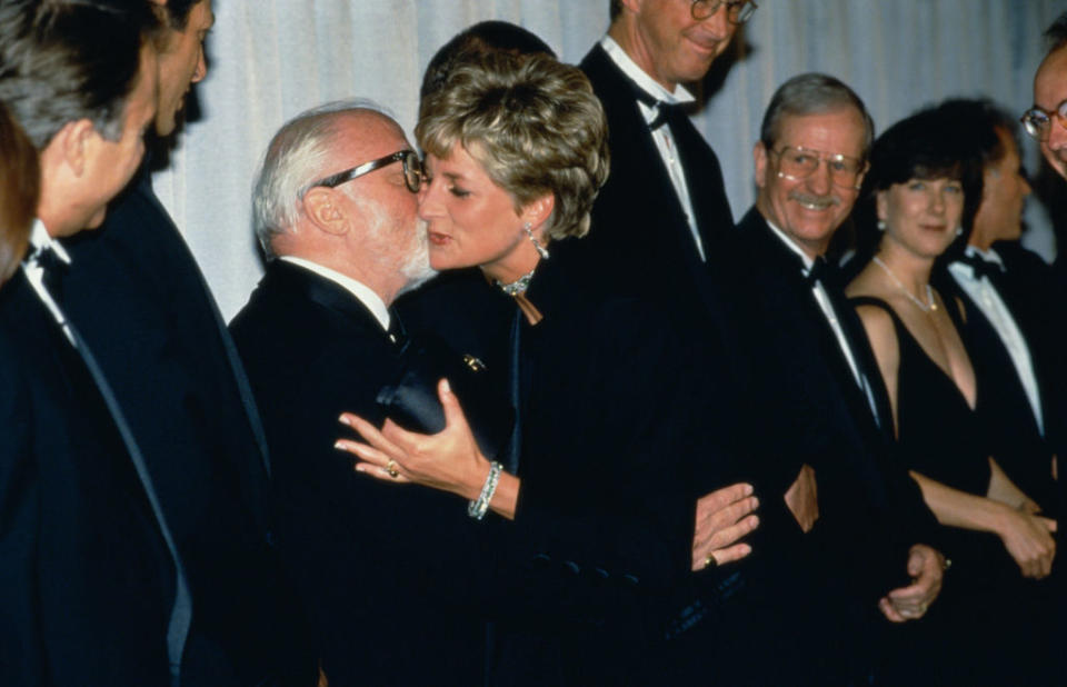 I don't know who the people in the image are. A woman in an elegant dress kisses a man on the cheek while others in formal attire stand in the background