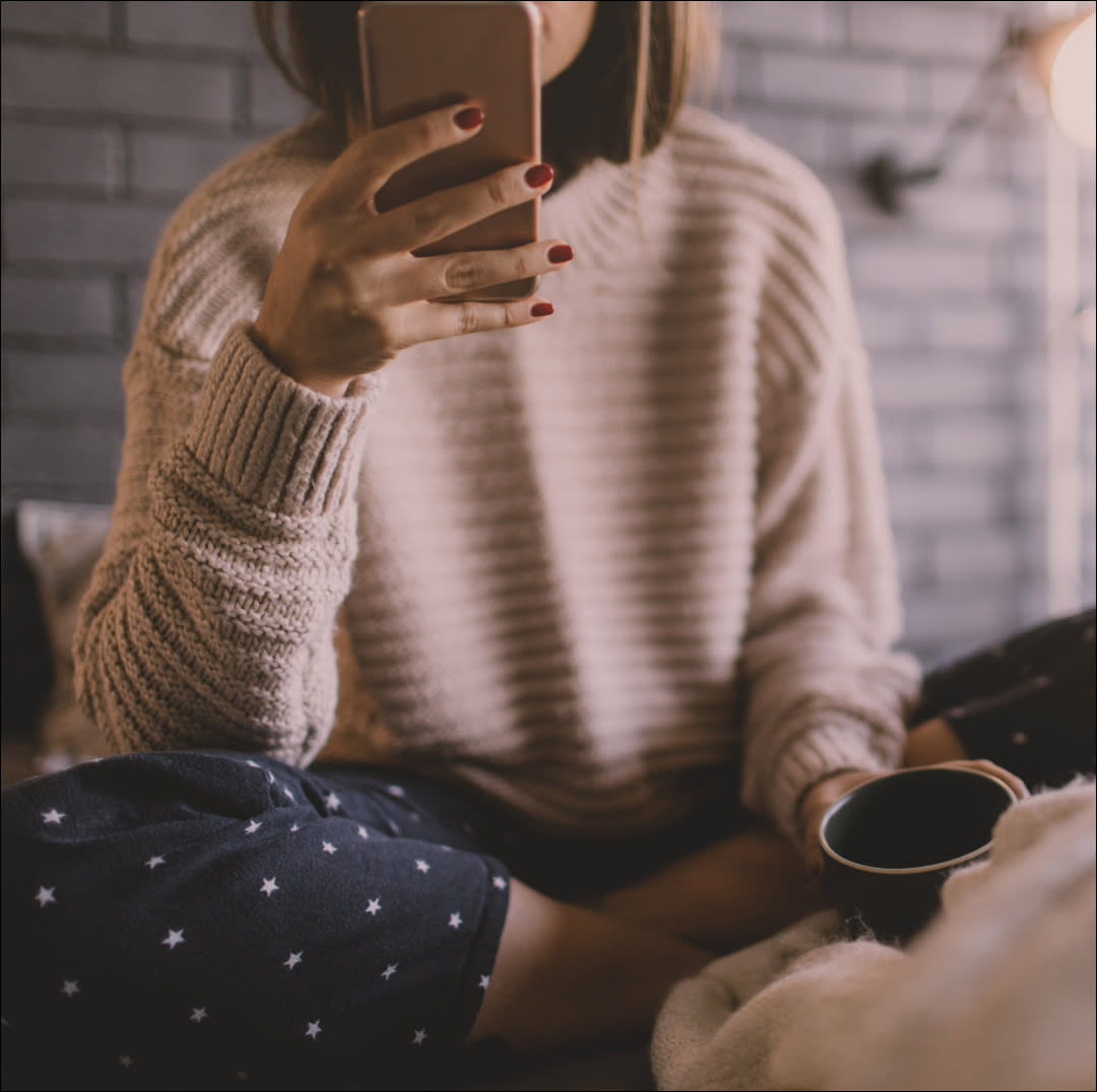  Photo of smiling young woman drinking first morning coffee in her bed, and reading online news on a smart phone. 