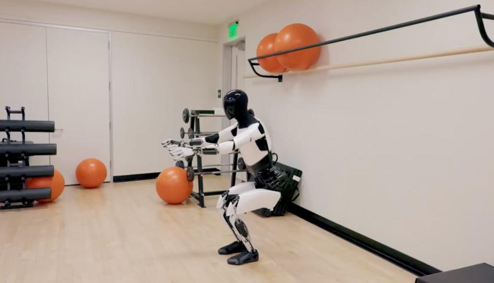 A still image from Tesla's promotional video of its robot Optimus where it is doing a squat with its arms extended in a gym