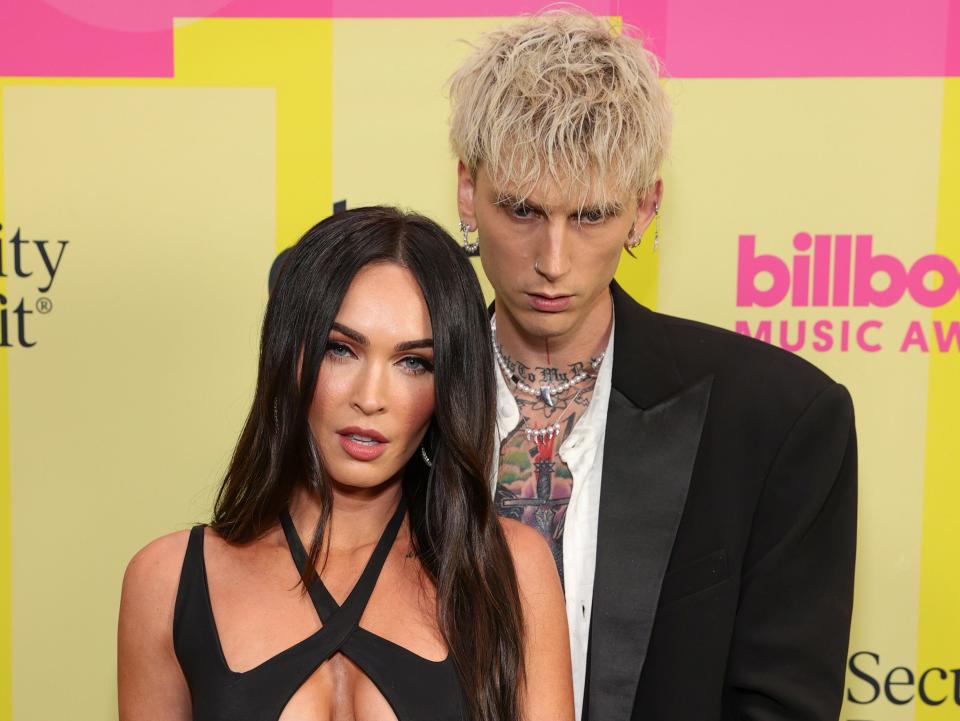megan fox in a black strappy dress and machine gun kelly in a black suit with an open white button down standing in front of a yellow and pink background