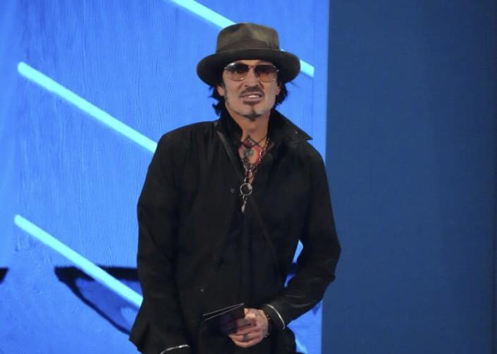 A man in dark sunglasses and a hat presents an award onstage