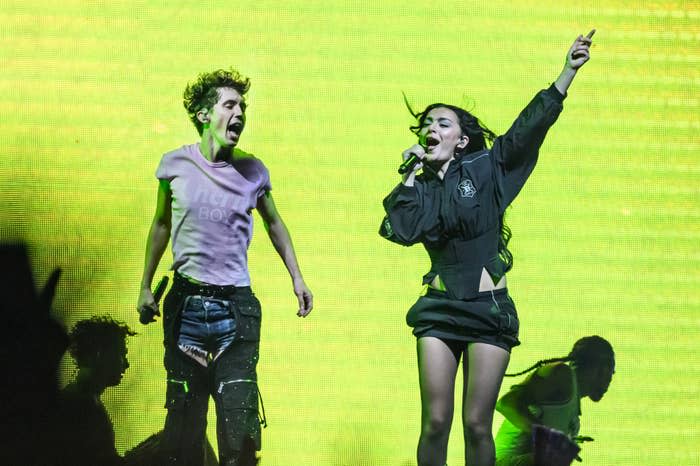 Troye Sivan and Charli XCX performing energetically on stage against a bright background. Troye wears a casual outfit; Charli wears a sporty ensemble