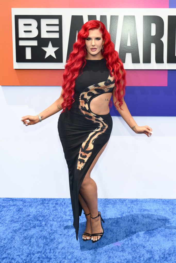 Justina Valentine poses on the BET Awards red carpet in a sleek black dress with a cut-out section and a high slit. She has long wavy hair and is wearing black heels