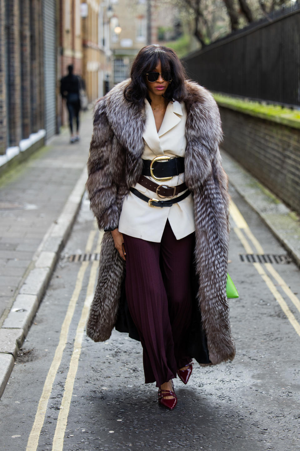 A woman wears three black belts over a white blazer and red pants with a fur coat.