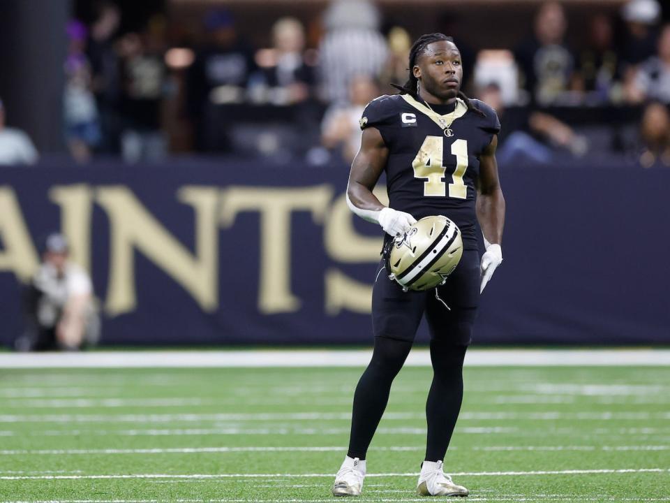 Alvin Kamara stands on the field and holds his helmet.