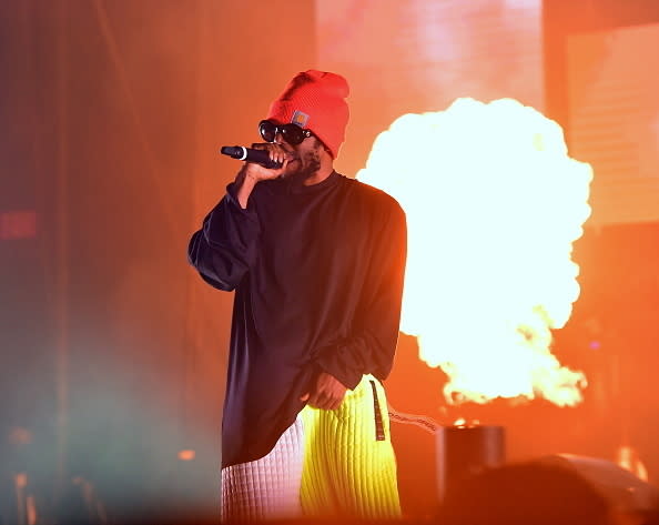 Man performing on stage with microphone, flame effect in background, wearing a beanie and long-sleeve shirt