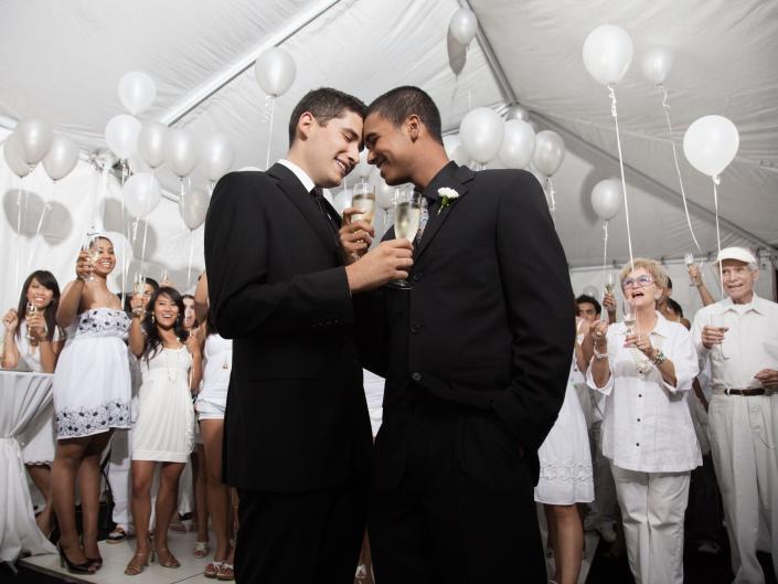 Couple standing in suits in the middle of a wedding reception in a white tent with balloons