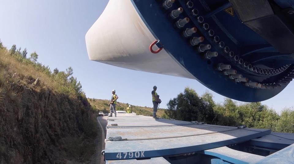 Two workers on the back of a truck transporting a section of a wind turbine.