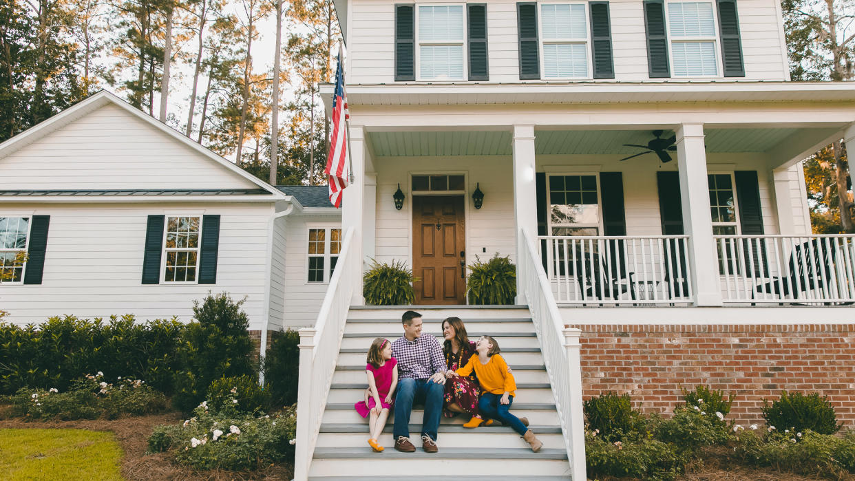 Family sitting outside on the steps of a new construction white siding farmhouse in the suburbs.