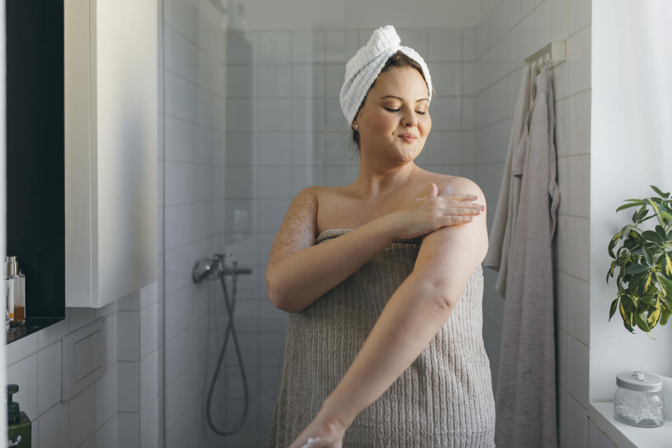 A woman stands in a bathroom with a towel wrapped around her body and another on her head, gently applying lotion to her arm, creating a self-care ritual
