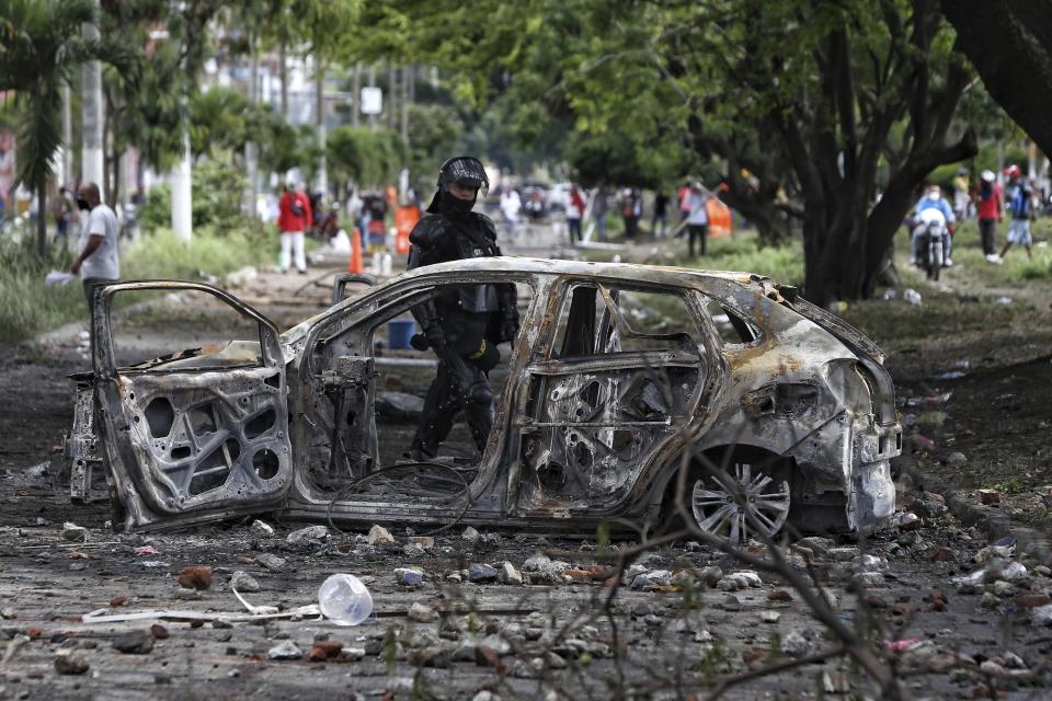 A police officer in riot gear walks past the charred remains of a car that was burned by protesters after a night of clashes with police in Yumbo, near Cali, Colombia, Tuesday, May 18, 2021. Colombians have taken to the streets for weeks across the country after the government proposed tax increases on public services, fuel, wages and pensions, but have continued even after President Ivan Duque walked back the tax hike. (AP Photo/Andres Gonzalez)