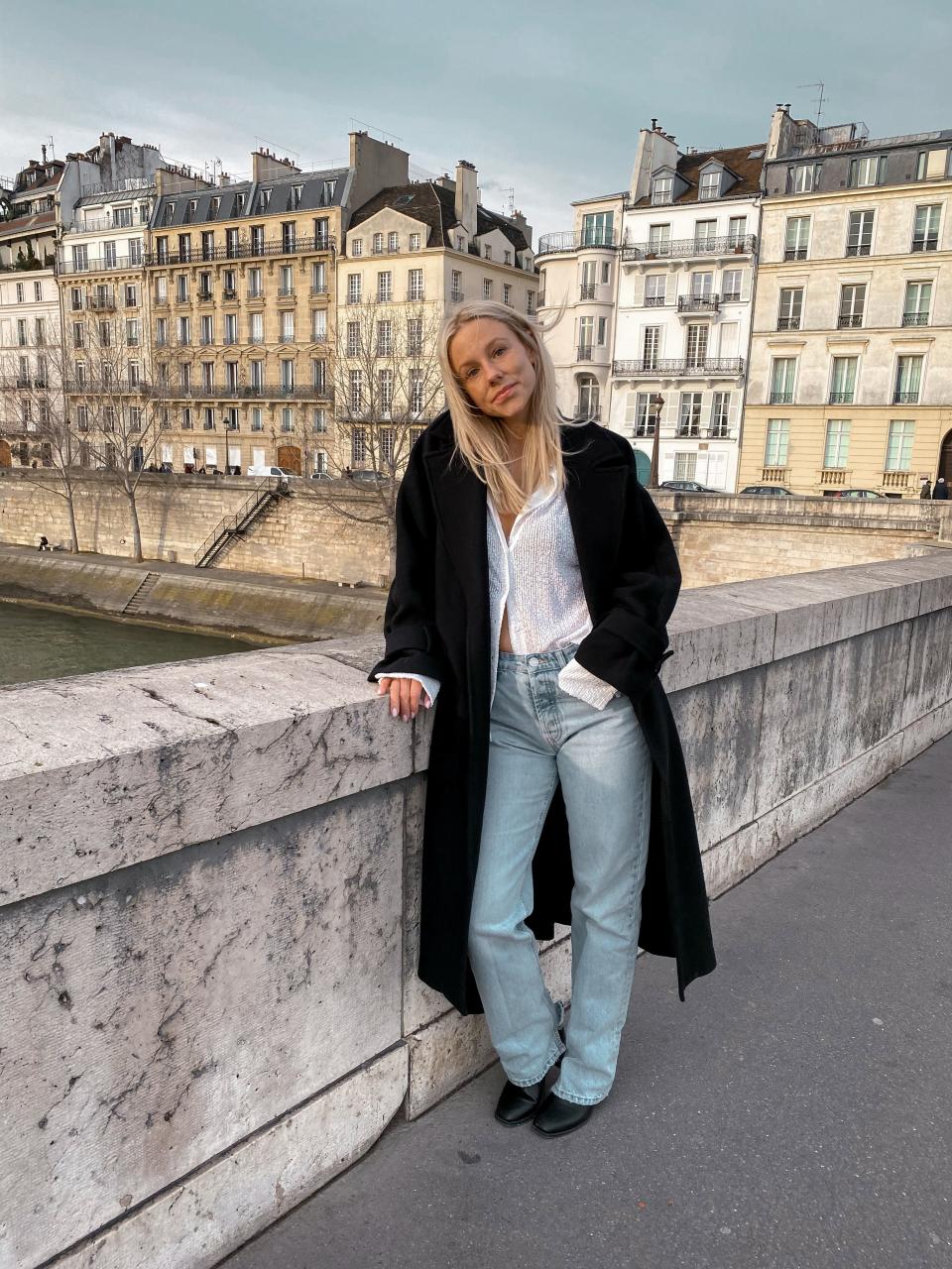 A woman with long blonde hair posing next to a bridge while wearing light blue jeans and a white shirt with a large black overcoat.