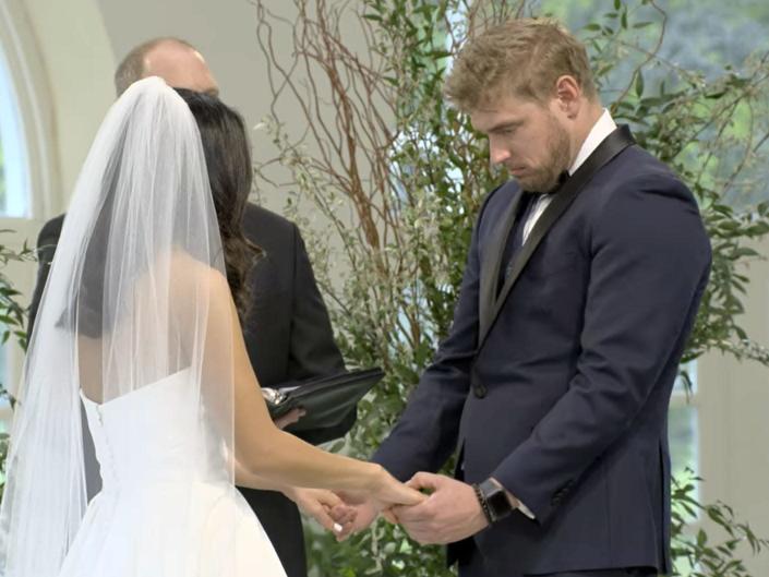 A woman wearing a white strapless dress a veil stands in front of a man in a navy tuxedo at an altar.