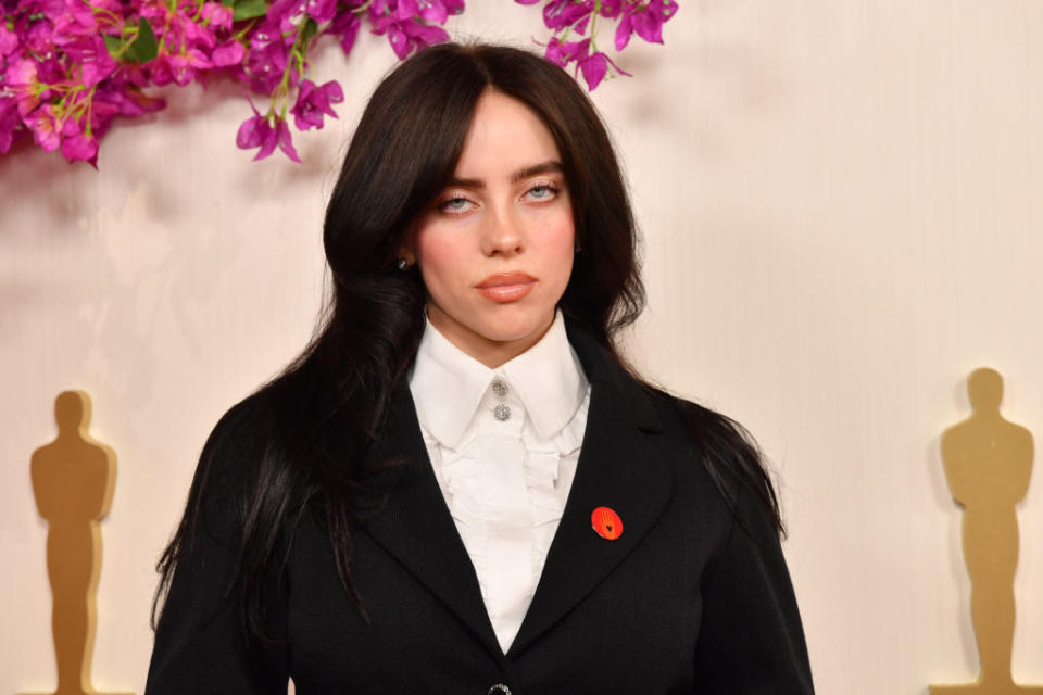 Billie Eilish in a white shirt and black blazer at an event with Oscar statues in the background