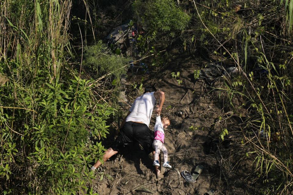 A man holds a child by their arm up as he climbs a muddy a river bank