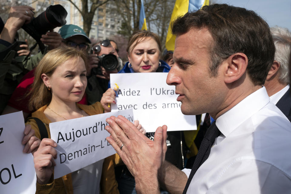 Emmanuel Macron brings his hands together as he speaks to protesters holding signs saying: Help Ukraine with arms, and Today Kyiv, Tomorrow [not visible, but possibly Europe].