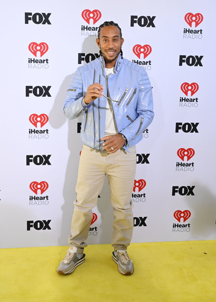 Ludacris at an event in a light jacket and sneakers, posing with one hand up, smiling